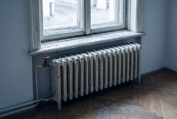 An old cast-iron radiator under a timber window frame with peeling paint, on a worn parquet floor